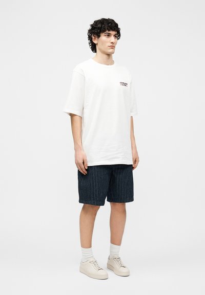 Νέος άνδρας που στέκεται, φορώντας ένα λευκό υπερμεγέθη μπλουζάκι με μικρό κείμενο στο στήθος, σκούρα μπλε σορτς με ρίγες, λευκές κάλτσες και λευκά αθλητικά παπούτσια.