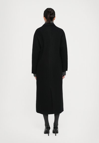 Femme de dos, portant un long manteau noir, des collants gris et des chaussures à talons noirs, devant un fond blanc uni.