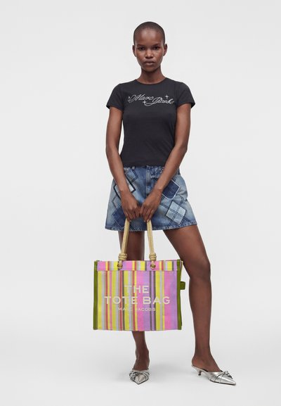 Femme debout tenant un grand sac cabas rayé, porte un t-shirt noir, une mini-jupe en jean et des escarpins argentés pointus sur fond uni.