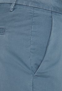Gros plan sur une poche de pantalon bleu clair avec des coutures visibles et une passant de ceinture sur une texture de tissu.