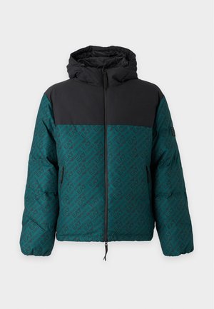 Jeune homme portant une veste vert foncé et noire ainsi qu'un pantalon noir, tenant un sac fourre-tout noir dans une main, regardant sur le côté.