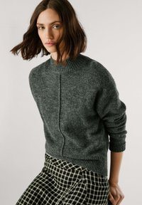 Sweater de malha cinza escuro com gola alta, punhos canelados e detalhes de costura subtis, combinado com uma saia xadrez em preto e branco.