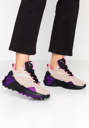 Person wearing beige, black, purple, and pink sporty sneakers with black laces and black pants, standing on a white floor.