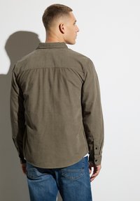 Olive green button-up shirt with long sleeves, rounded hem, and textured fabric. Paired with blue jeans. Back view shows seam details.