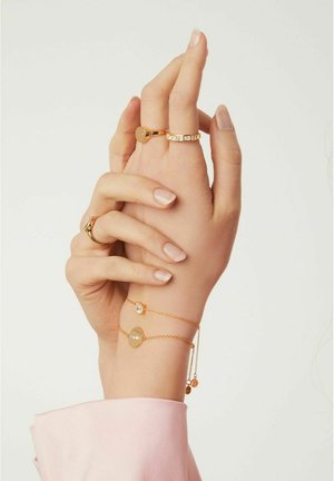 Guldtonade ringar och armband på händer, med cirkulära berlocker och en glittrande sten. Slät yta med eleganta kedjedetaljer.