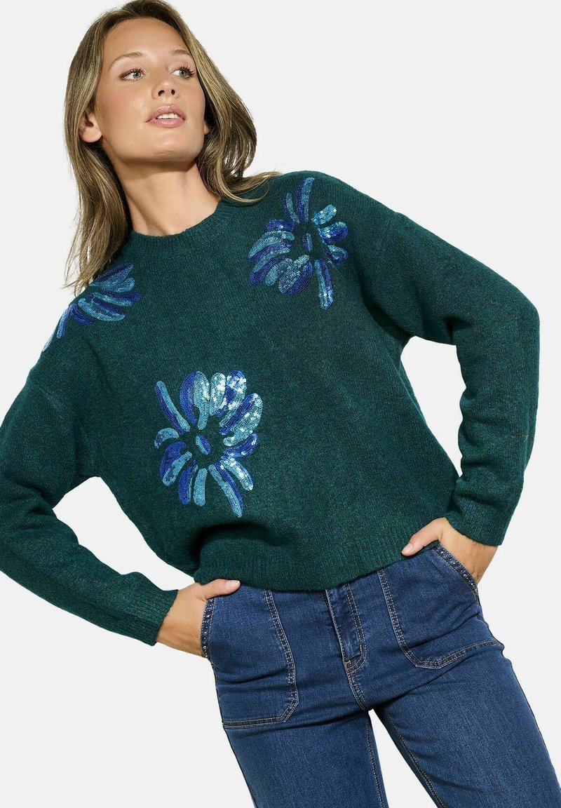 Maglione lavorato a maglia verde con motivi floreali in paillettes blu sul davanti. Collo tondo e maniche lunghe; abbinato a jeans in denim.