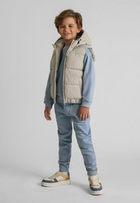 Beige vadderad väst med huva, ljusblå sweatshirt, matchande träningsbyxor och sneakers med beige, blå och vita detaljer.