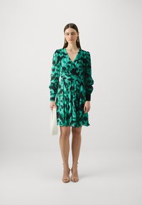 Robe portefeuille verte avec un motif noir et turquoise, manches longues, ceinture à la taille et ourlet fluide. Associée à des talons beiges à lanières et un sac blanc.