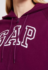 Gros plan sur une personne portant un sweat à capuche zippé violet de GAP avec des lettres blanches en contour et des cordons visibles.