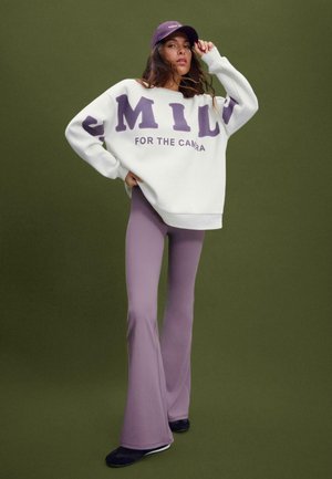 Felpa bianca con scritte viola, "SORRIDI PER LA MACCHINA", abbinata a pantaloni viola a gamba tapered e un cappellino coordinato. Le scarpe nere completano il look.