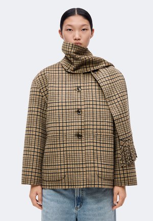 Femme portant un manteau à carreaux beige et noir boutonné avec une écharpe assortie enroulée autour du cou, assorti à un jean bleu clair.