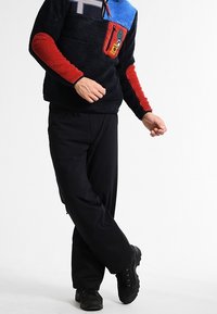 Pullover in pile con pannelli multicolori: corpo blu navy, maniche rosse, colletto blu. Presenta una tasca con zip e un dettaglio del logo. Pantaloni neri e stivali.