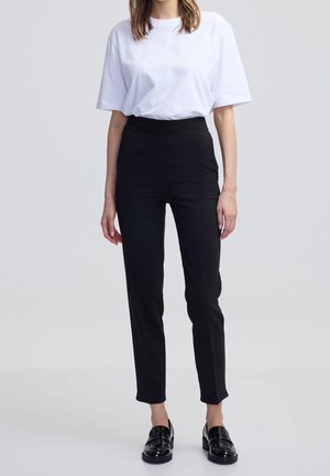 Persoon die een effen wit T-shirt, slanke zwarte broek en zwarte loafers draagt, staand voor een lichtgrijze achtergrond.
