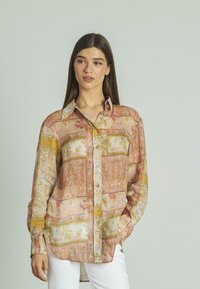 Camisa ligera, con un patrón en rosa y amarillo, cuello con solapa, botón en la parte delantera y mangas largas. La tela tiene una textura suave y transparente.