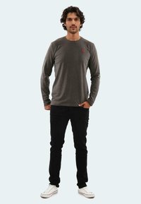 Graues Longsleeve-Shirt mit rundem Ausschnitt, kombiniert mit schwarzen Hosen und weißen Turnschuhen. Hat ein kleines rotes Logo auf der Brust.