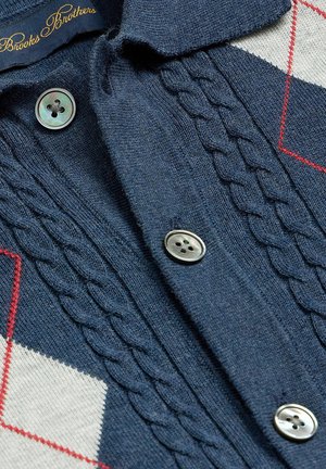 Granatowy sweter z dzianiny z wzorem w warkocze, srebrnymi guzikami z czterema dziurkami oraz białymi i czerwonymi akcentami w romby po bokach, oznaczony metką Brooks Brothers.