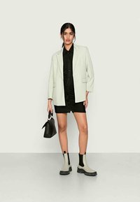 Blazer oversized verde claro, camisa preta de botões, vestido mini franzido preto, botas cinzas até ao tornozelo com detalhes pretos e uma pequena bolsa preta.