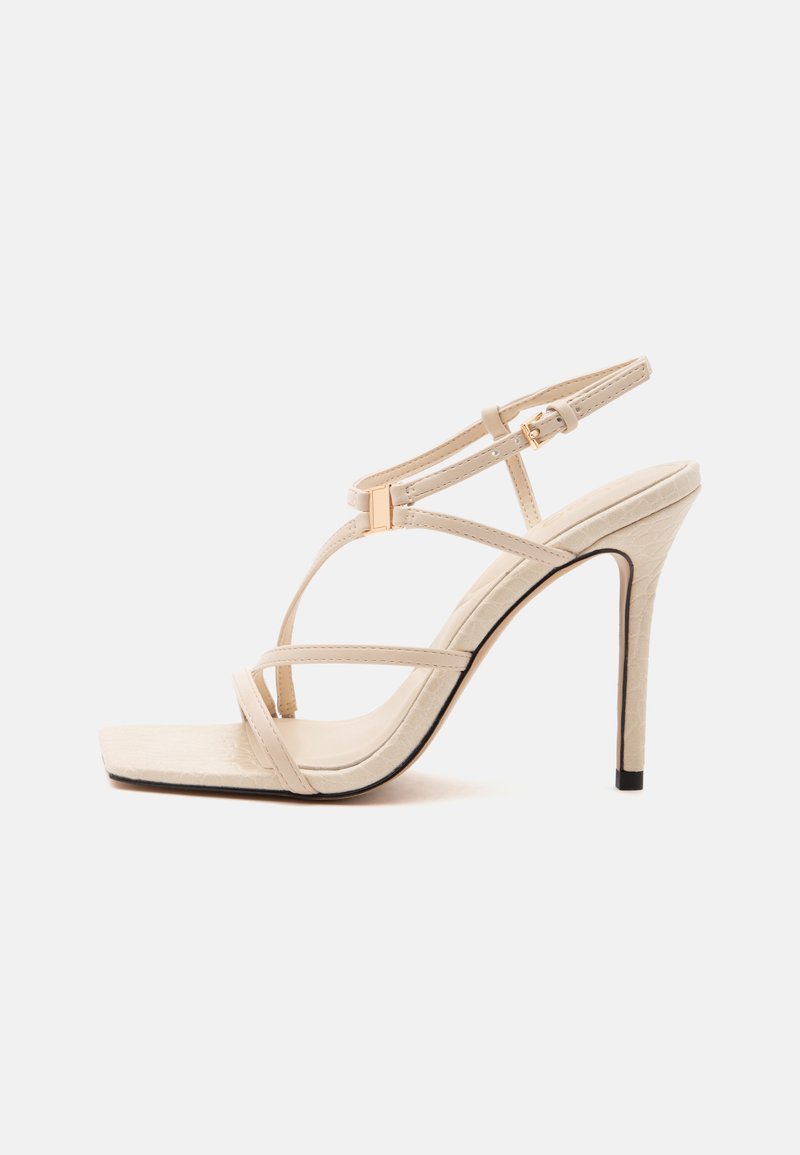 Sandale beige à talon haut avec fines lanières, bout carré, fermeture à boucle et talon stilettos sur fond blanc.