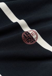 Tissu sombre avec une large bande verticale blanche et un petit logo rond contenant des lettres de couleur brun rougeâtre près de la bande.