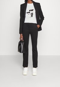 Svart kavaj över en vit t-shirt med grafik, matchat med svarta byxor och vita sneakers. Håller en svart handväska.