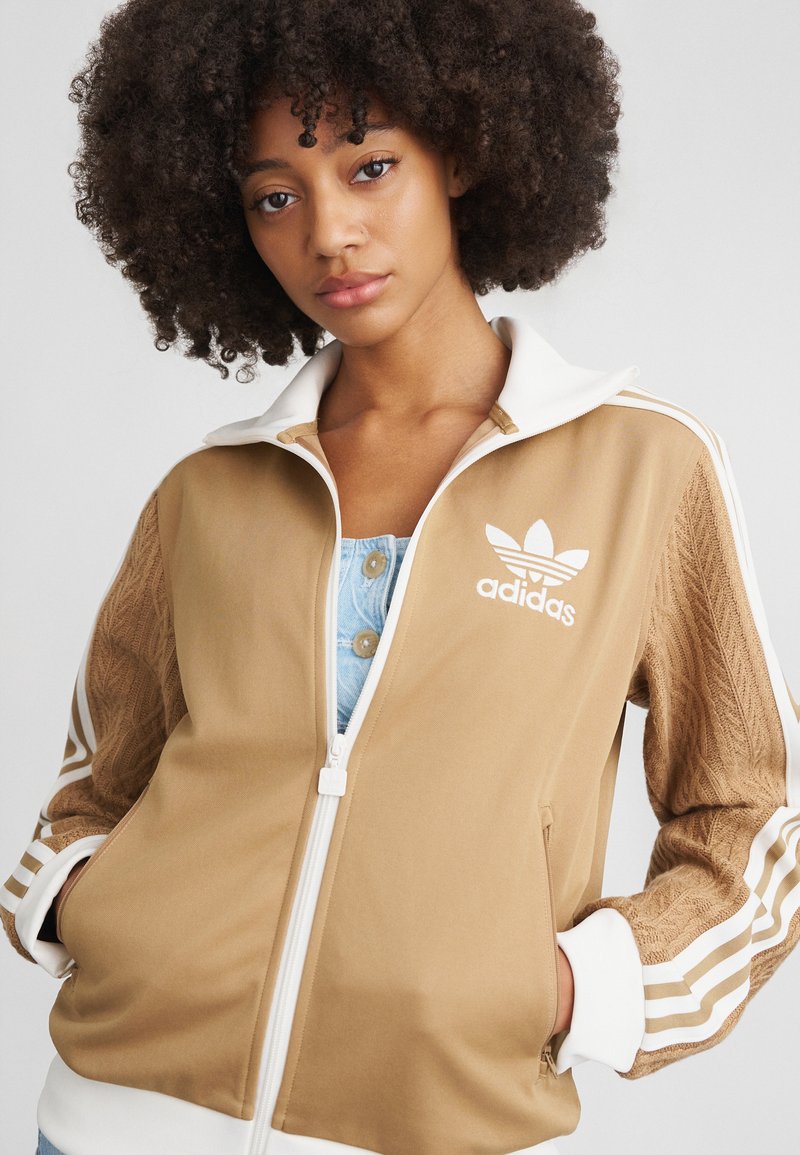 Braune Sportjacke mit einem weißen Reißverschluss und Kragen. Verfügt über strukturierte braune Ärmel und drei weiße Streifen an jedem Ärmel. Adidas-Logo auf der Brust.