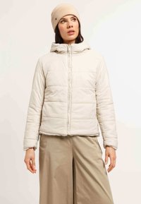 Donna che indossa un berretto beige e una leggera giacca a vento con cappuccio, abbinata a pantaloni beige larghi, che guarda leggermente verso l'alto a destra.