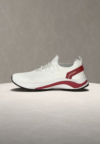 Chaussure de sport blanche avec des accents rouges, tige en mesh, fermeture à lacets, et semelle épaisse et rembourrée sur une surface et un fond neutres.