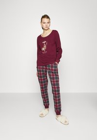 Bordeaux kleurige lange mouwen top met bloemenprint, gecombineerd met grijze en rode geruite broek en crèmekleurige, fluffy slippers. Casual, relaxte pasvorm.