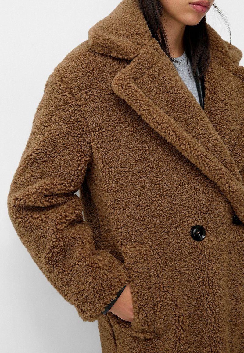 Femme portant un manteau teddy texturé marron avec des boutons noirs, une main dans sa poche, sur un fond clair uni.