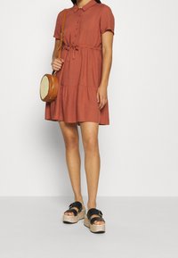 Robe à manches courtes, boutonnée, de couleur rouille, avec une taille cintrée et une jupe à volants. Assortie à des sandales à plateforme noires et un sac rond.