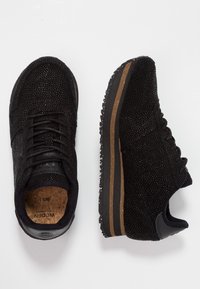 Černé tenisky s texturovaným povrchem, vybavené korkovou stélkou, gumovou podrážkou a hnědým akcentem podél okraje podrážky. Design na šněrování.