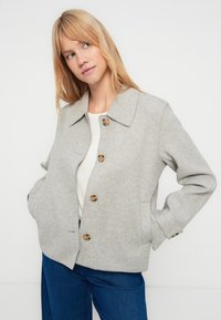 Giacca grigia in misto lana con colletto, tre bottoni marroni e tasche laterali, abbinata a una maglia bianca e jeans blu.