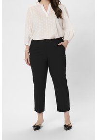 Mujer que lleva una blusa blanca con textura y mangas tres cuartos, pantalones negros capri, zapatos planos negros de punta y una pulsera de perlas.