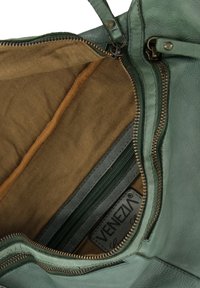 Zelená kožená kabelka uvnitř s hnědou textilní podšívkou, více zipovými přihrádkami a viditelnou logovou cedulkou na boku.