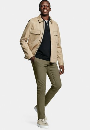 Lachende man staand met handen in de zakken, draagt een beige jas, zwarte trui met rits, olijfgroene broek en beige sneakers op witte achtergrond.