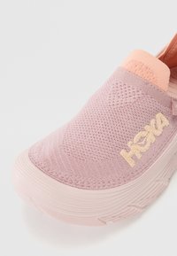 Sandală roz, ușor de încălțat, cu un superior din tricot texturat, având un detaliu în nuanță piersică la toc și o talpă din cauciuc de un roz deschis. Logo-ul HOKA este prezent.