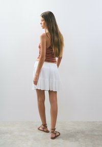 Top de tirantes de ganchillo color oliva y falda corta blanca con volantes. La modelo lleva sandalias marrones con tiras. Se puede ver su cabello largo y liso desde atrás.