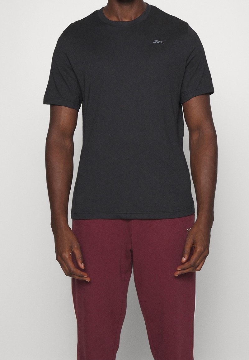 T-shirt nera a maniche corte realizzata in tessuto morbido, con scollo tondo e un piccolo logo sulla parte superiore sinistra. Indossata con pantaloni tuta bordeaux.