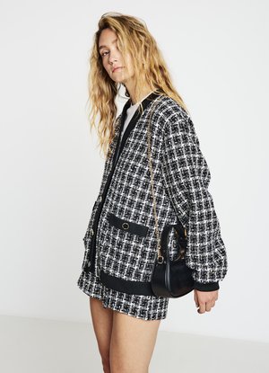 Giacca a quadri bianca e nera dal taglio comodo, con due tasche e polsini a costine. Una piccola borsa nera è tenuta in una mano.