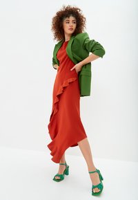 Rode jurk met een gerimpelde zoom, gecombineerd met een groene blazer. Groene sandaaltjes met hoge hakken maken de outfit compleet. De achtergrond is helder wit.