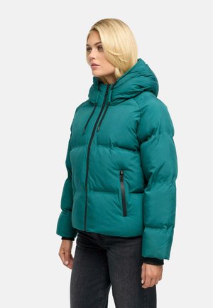 Femme portant une doudoune bleu sarcelle avec une capuche et des poches zippées noires, debout de profil sur un fond uni.