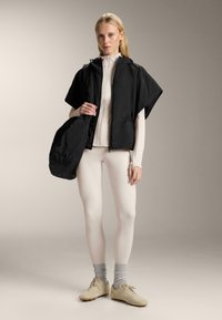 Zwart cropped vest met capuchon en trekkoord, gedragen over een licht shirt en crème legging. Beige schoeisel en grijze sokken maken de outfit compleet.