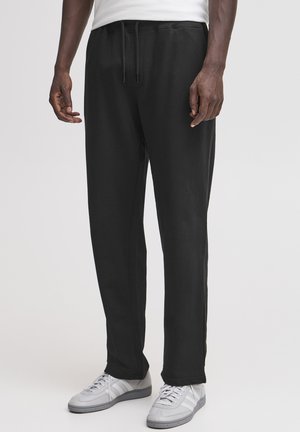 Pantaloni della tuta neri in tessuto morbido con fascia elastica in vita, coulisse regolabile e tasche laterali. Abbinati a scarpe da ginnastica grigio chiaro.