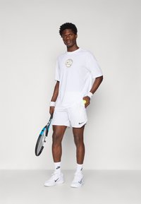 Hvit sportst-skjorte med gulllogo, hvite shorts, tennisracket i hånden, hvite joggesko, håndleddbånd. Jevn tekstur, atletisk passform.
