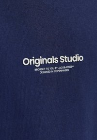Sötétkék pamut póló, fehér felirattal: "Az Originals Stúdiót a JACK&JONES® hozta létre. Tervezve Koppenhágában."