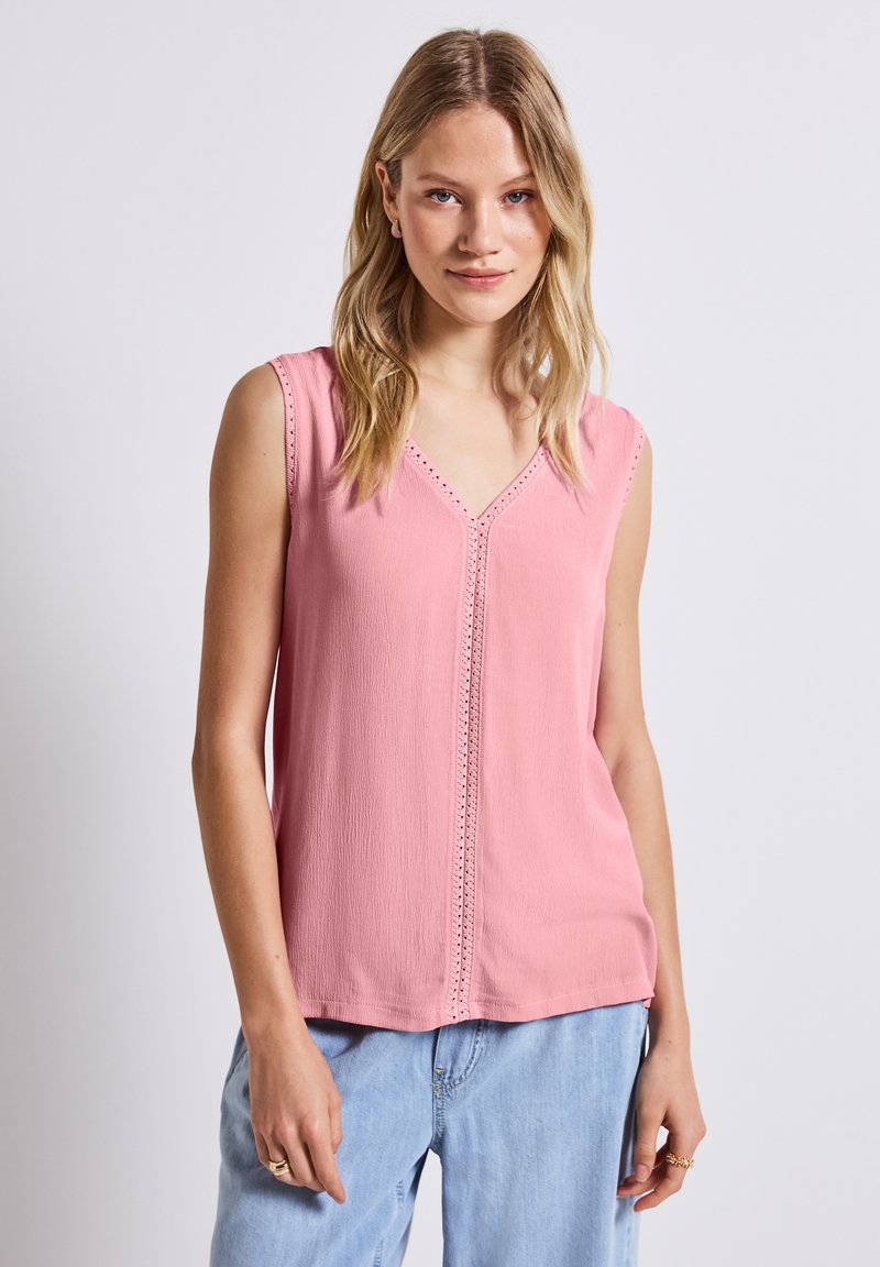 Blouse sans manches rose avec un col en V, ornée de dentelle le long des bords et d'un tissu texturé. Associée à un jeans bleu clair.