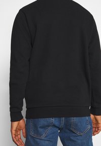 Persona vestida con una camiseta negra de manga larga y jeans de mezclilla azul, vista desde detrás, contra un fondo blanco liso.