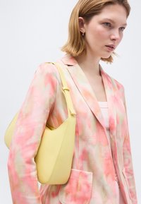 Νεαρή γυναίκα που φοράει σακάκι με παστέλ ροζ και πορτοκαλί σχέδια, κρατώντας ανοιχτό κίτρινο τσαντάκι ώμου, με κοντά ξανθά μαλλιά και χρυσούς κρίκους σκουλαρίκια.