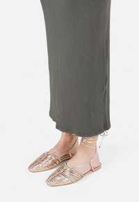 Metalne rosegold sandale s ravnim potplatom, tkanim dekorom, odprtim prstom in naramnicami okoli gležnjev, skupaj z rebrastim olivno zelenim maksi oblekcem.