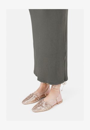 Fém rózsaarany lapos szandál, fonott dizájnnal, nyitott orral és bokapántokkal, olajzöld, bordázott maxi ruhával párosítva.